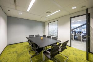 Modern conference room with a black table, eight chairs, and green carpet, connected to an open office area at Jacob Bontiusplaats.